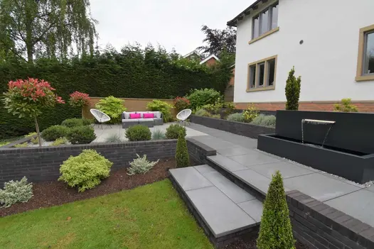 Patio across back of white house, with seating area, steps to lawn and black wall with water spout. Design by Creative Gardens.