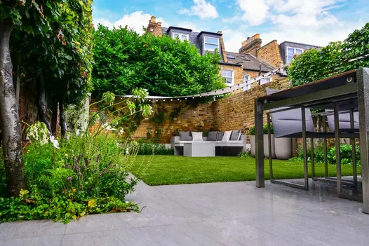 Walled back garden by Harrington Porter. Trendy black porcelain patio edging lawn with outdoor furniture. Verdant border on left.