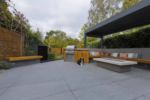 Trendy Black Porcelain paved outdoor seating and kitchen area with fire-table, covered by pergola. Surrounded by hedge and fence.