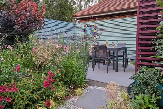 Raised patio area bordered on three sides by horizontal slatted screens with lush planting and a table and chairs.
