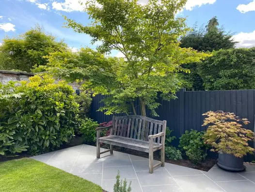 Under a tree, a bench sits on Trendy Black Porcelain paving. Border with shrubs and dark fence behind. By Dee Stewart Gardens.
