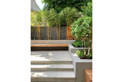 Urban Grey porcelain steps with downstand rise to matching paved area with cedar bench, edged by trees in raised beds.