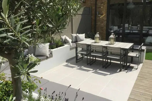 Cushioned built-in benches edge Urban Grey Porcelain patio with dining set. Part of tree in foreground. Lanterns on table.