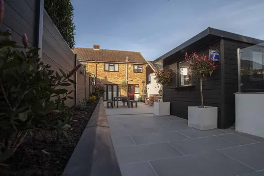Long raised bed edged with twice-weathered coping runs down length of garden completely paved in Urban Grey porcelain paving.