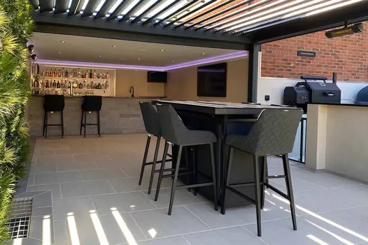 High table with stools under louvred pergola. Covered outdoor bar area in background. All paved in Urban Grey Porcelain.