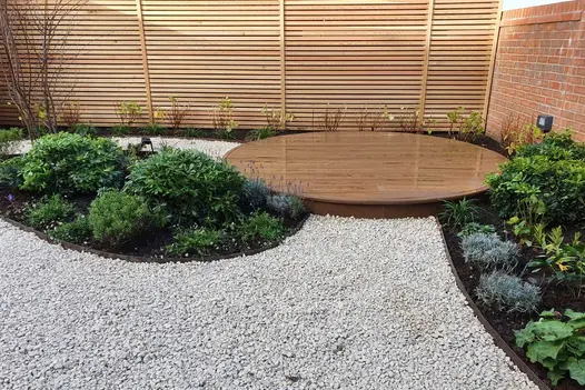 Round raised Millboard decking finished with flexible bullnose edging in corner of gravel-paved garden with curve-edged beds.