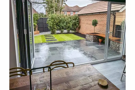 View over wooden table through open bifold doors to garden with Brazilian black slate patio. Slabs set into lawn lead to gravel.