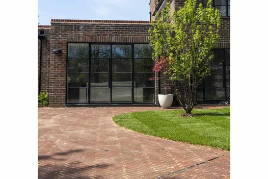 Low extension with 4 panels of glass door overlooks driveway of Abbey Dark Multi clay pavers, curving around lawn with tree.