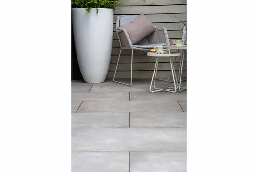 Tall white planter sits on Venetian Grey Porcelain Paving against slatted fence, next to metal and rattan chair and low tables.