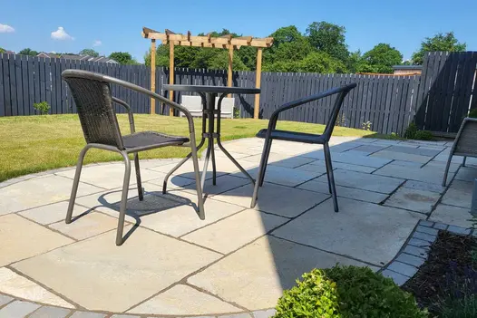 Slim table and 2 metal chairs on patio of Antique Yellow limestone paving, with setts along curved edge. Lawn with pergola beyond.
