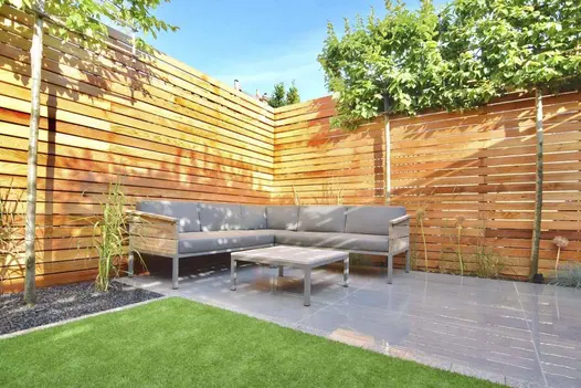 Outdoor corner sofa and table sits on Trendy Black vitrified porcelain patio, edged by tall fence and box-head hornbeams.