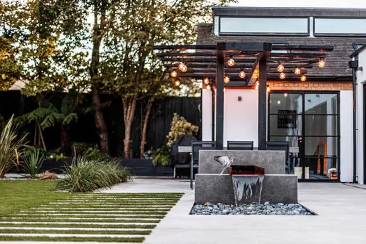 Pigeon lands on waterfeature clad in Vulcano Ceniza Luxury Designclad, wooden pergola above adorned in string lights.