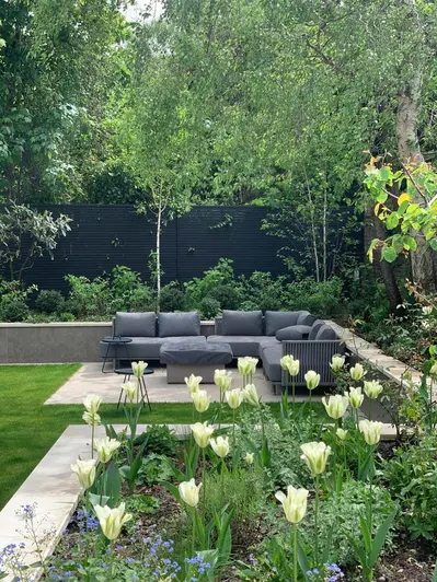 Patio in corner of garden with shrubs and trees in border edged by low wall faced with Vulcano Ceniza porcelain wall cladding.