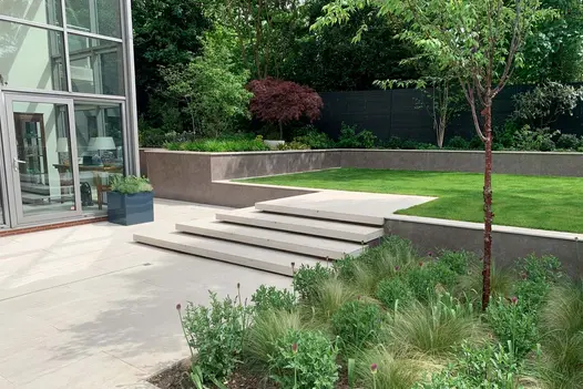Light coloured patio and matching steps lead up to lawn with raised flowerbeds surrounding, clad in vulcano ceniza designclad.
