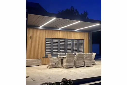 Outdoor dining area under lit up roof has warm teak composite battens cladding the building next to it..