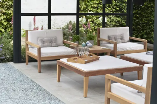 Rectilinear wood-framed seats and table sit on Jura Grey limestone contemporary paving under pergola with planted beds behind. 