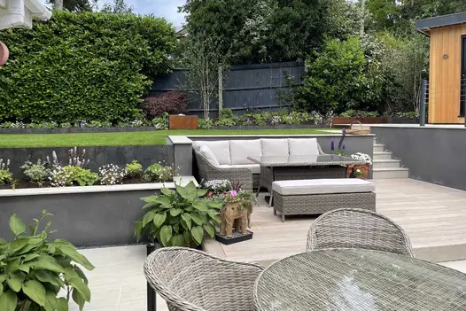 Aerial view of an elevated lawn area descending to a cinnamon designboard composite decking space with a corner rattan couch and glass table.