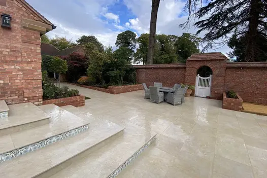 5 wide steps in Golden Stone porcelain patio tiles and patterned risers descend to paved area with tall brick wall boundary. 