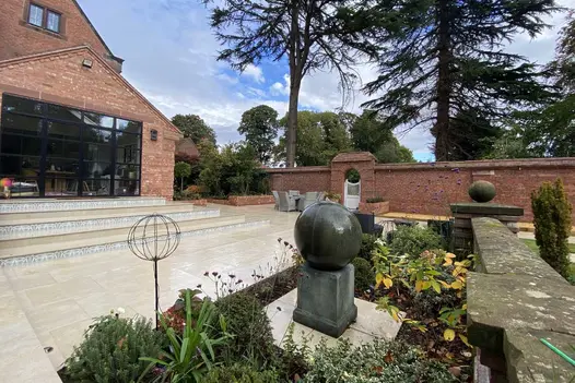 Large patio of 900x600 Golden Stone porcelain paving wrapped around corner of house, with brick raised beds and water feature. 